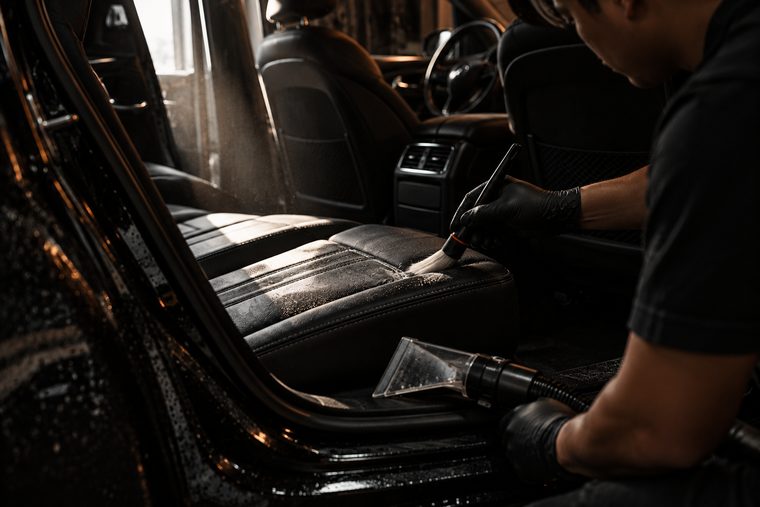 A gloved hand working a detailing brush into the stitching of a black leather seat, shot through the open rear door.