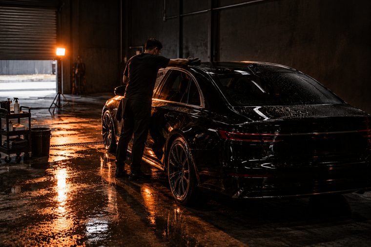 A detailer leaning across the roof of a half-dried black car with a microfibre towel, alone in the lit bay.